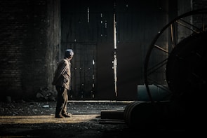 A solitary figure stands in a dimly lit industrial space, gazing towards a crack in a large, rusty door. The light seeping through creates a stark contrast against the dark interior, illuminating the person and casting long shadows. The scene presents an atmosphere of mystery and introspection.