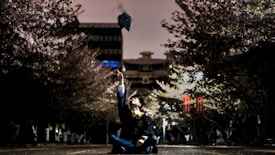 A person wearing a graduation gown sits on the ground, joyously tossing a graduation cap into the air. The scene is framed by cherry blossom trees on either side, creating a dramatic and celebratory atmosphere. In the background, there are buildings and illuminated banners.
