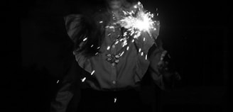 A person holding a lit sparkler in a dark environment, with a shirt featuring ruffled details and a brooch. The sparkler emits bright sparkles that contrast sharply with the surrounding darkness.