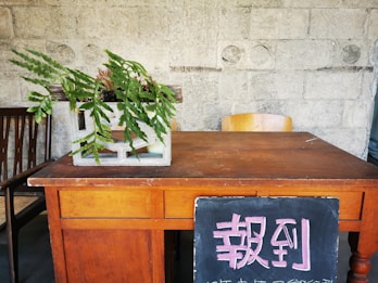 A wooden desk is positioned against a textured concrete wall with circular imprints. On the desk sits a potted plant with lush green leaves, housed in a concrete block style planter. There's a wooden chair behind the desk, and another chair partially visible to the side. A chalkboard with bold pink Chinese characters leans against the front of the desk.