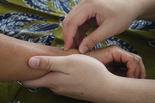 Two hands gently hold and press the wrist of another person, suggesting a massage or therapeutic touch. The background features a patterned fabric with blue and green tones.
