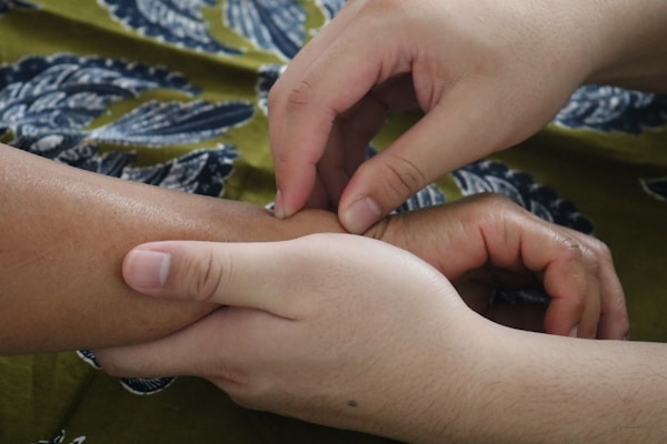Two hands gently hold and press the wrist of another person, suggesting a massage or therapeutic touch. The background features a patterned fabric with blue and green tones.
