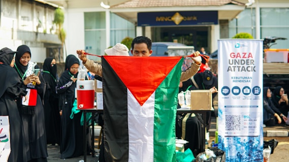 A group of people, some wearing black clothing with green accents, are gathered outside. One person holds a Palestinian flag prominently. A banner next to them reads 'Gaza Under Attack' with additional information.