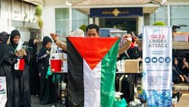 A group of people, some wearing black clothing with green accents, are gathered outside. One person holds a Palestinian flag prominently. A banner next to them reads 'Gaza Under Attack' with additional information.