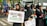 Several people wearing black outfits and headscarves stand together, holding signs supporting Palestine. One sign reads 'I Stand With Palestine,' while another promotes purchasing something for a cause. Some individuals have small Palestinian flags pinned to their clothing.