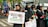Several people wearing black outfits and headscarves stand together, holding signs supporting Palestine. One sign reads 'I Stand With Palestine,' while another promotes purchasing something for a cause. Some individuals have small Palestinian flags pinned to their clothing.