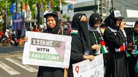 Several people wearing black outfits and headscarves stand together, holding signs supporting Palestine. One sign reads 'I Stand With Palestine,' while another promotes purchasing something for a cause. Some individuals have small Palestinian flags pinned to their clothing.