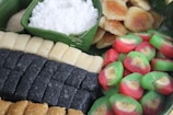 A variety of colorful traditional desserts are arranged on a banana leaf. Rectangular and square-shaped treats are predominantly black, beige, and dark brown, alongside circular snacks in vibrant red and green hues. A portion of shredded white coconut is placed in a green container made from a leaf.