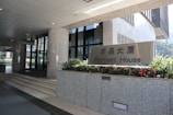 Entrance to an office building with a sign for Fairmont House. The area is clean and modern, featuring granite pillars, glass doors, and flower beds with vibrant red flowers.