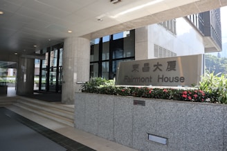 Entrance to an office building with a sign for Fairmont House. The area is clean and modern, featuring granite pillars, glass doors, and flower beds with vibrant red flowers.
