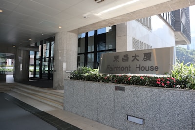 Entrance to an office building with a sign for Fairmont House. The area is clean and modern, featuring granite pillars, glass doors, and flower beds with vibrant red flowers.