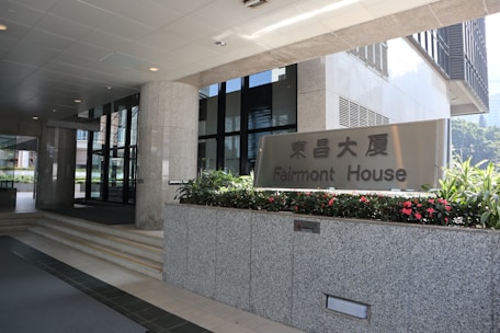Entrance to an office building with a sign for Fairmont House. The area is clean and modern, featuring granite pillars, glass doors, and flower beds with vibrant red flowers.