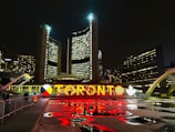 A nighttime cityscape featuring a large, illuminated sign displaying the name Toronto in vibrant letters. The backdrop includes towering buildings with numerous windows, and there are reflections of the lights on the wet ground. Some people are walking by, and the surrounding architecture is modern and distinctive.