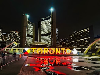 A nighttime cityscape featuring a large, illuminated sign displaying the name Toronto in vibrant letters. The backdrop includes towering buildings with numerous windows, and there are reflections of the lights on the wet ground. Some people are walking by, and the surrounding architecture is modern and distinctive.