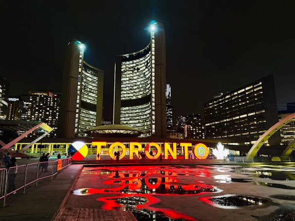 A nighttime cityscape featuring a large, illuminated sign displaying the name Toronto in vibrant letters. The backdrop includes towering buildings with numerous windows, and there are reflections of the lights on the wet ground. Some people are walking by, and the surrounding architecture is modern and distinctive.
