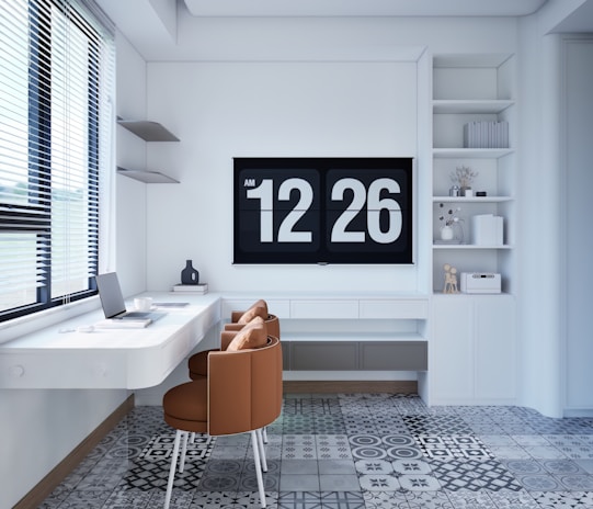 A modern, minimalist home office setup features a floating white desk with a laptop, a coffee cup, and a small decorative object. Two brown chairs provide seating. The back wall is adorned with a large digital clock displaying the time 12:26. Shelving to the right holds books and small decor items. The floor is covered in patterned tiles, and a window with blinds allows natural light to brighten the room.