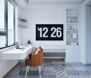 A modern, minimalist home office setup features a floating white desk with a laptop, a coffee cup, and a small decorative object. Two brown chairs provide seating. The back wall is adorned with a large digital clock displaying the time 12:26. Shelving to the right holds books and small decor items. The floor is covered in patterned tiles, and a window with blinds allows natural light to brighten the room.