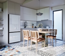 A modern kitchen with white cabinetry and a tiled backsplash. The floor is covered in patterned tiles, and there is a dining area with a rectangular table and four wooden chairs. A vase with flowers is placed on the table, and there is a pendant light hanging above. The kitchen includes a refrigerator, shelves with books, and some countertop items such as a black sink, cutting board, and additional plants. Natural light enters through a glass door on the right side.