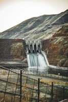A large dam structure with water flowing over the spillway surrounded by hills.