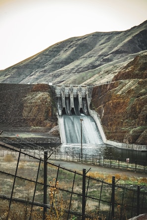 A large dam structure with water flowing over the spillway surrounded by hills.