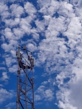 A tall metal telecommunications tower rises against a backdrop of a bright blue sky filled with scattered fluffy white clouds. The structure features antennas and equipment mounted at the top.