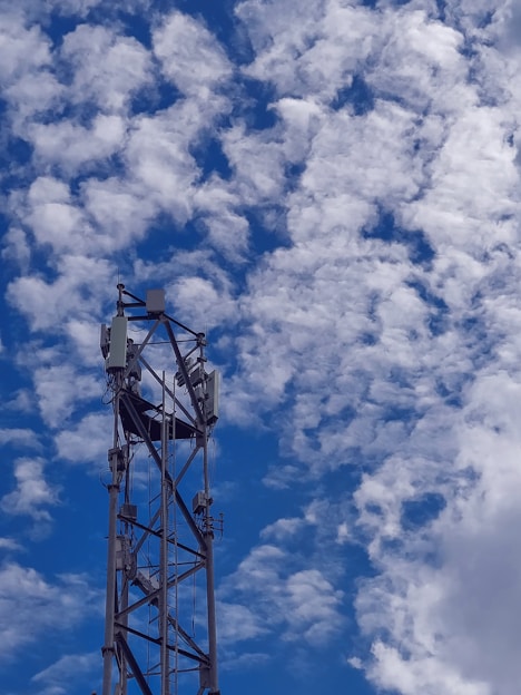A tall metal telecommunications tower rises against a backdrop of a bright blue sky filled with scattered fluffy white clouds. The structure features antennas and equipment mounted at the top.