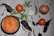 A flat lay of various ingredients includes a bowl of vibrant orange sauce, surrounded by fresh tomatoes, whole dried chilies, a head of garlic, a brown onion, and sprigs of cilantro. The background is a light wooden surface with scattered small red chilies.
