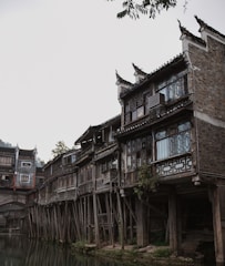 Traditional wooden stilt houses are built along a riverbank. The architecture features intricate wooden carvings and lattice windows. The scene has a rustic and historical feel, with lush greenery partially visible in the background and overcast skies above.