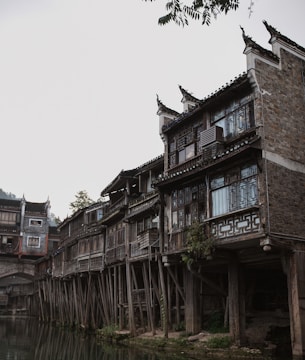 Traditional wooden stilt houses are built along a riverbank. The architecture features intricate wooden carvings and lattice windows. The scene has a rustic and historical feel, with lush greenery partially visible in the background and overcast skies above.