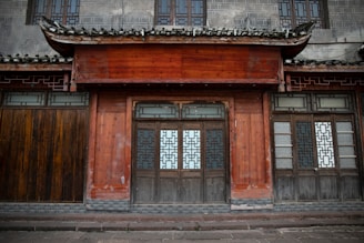 A traditional building with wooden doors and intricate geometric patterns on the windows. The exterior wall features a mix of aged wood and gray stone, topped with a tiled roof.