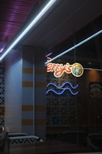 A dimly lit interior setting with a neon sign displaying the name of a business. The letters are partially visible, and there's a decorative wall with repetitive patterns. The scene includes tables and chairs arranged casually. Lighting is soft, and the ambiance appears relaxed.