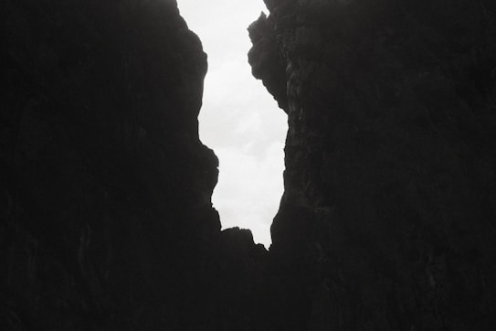 A narrow, irregularly shaped opening between two dark cliffs is visible, with a light sky in the background. The jagged edges of the rocks create a dramatic silhouette against the soft, overcast sky.