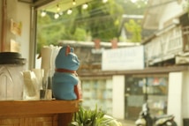 A wooden counter holds a variety of objects, including a blue ceramic cat figurine with a red collar, a blender, a spray bottle, and a glass jar with utensils. The background shows an outdoor scene through a window, featuring a blurred street view with buildings and green foliage.