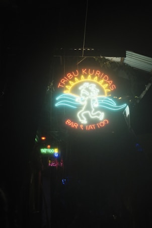 A neon sign with vibrant colors showcasing a sun with a face, along with decorative waves. The sign reads 'TRIBU KURIDAS BAR & TATTOO' and is positioned in a generally dark environment. Additional smaller neon signs are visible in the background, adding more colorful lighting to the scene.