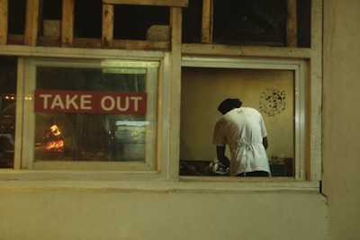 A person dressed in a white shirt and apron is seen from behind through a window labeled with a 'TAKE OUT' sign. The interior appears to be a kitchen with a wood-fired oven visible to the left, casting a warm glow.