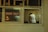 A person dressed in a white shirt and apron is seen from behind through a window labeled with a 'TAKE OUT' sign. The interior appears to be a kitchen with a wood-fired oven visible to the left, casting a warm glow.