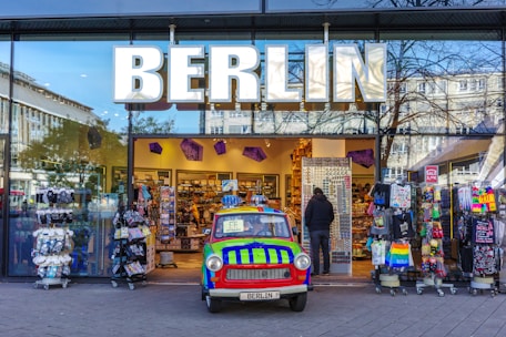 A colorful vintage car is parked in front of a Berlin souvenir shop. The shopfront features large, bold letters spelling 'BERLIN' above the entrance. Various souvenirs and merchandise are displayed on racks outside, including t-shirts and bags. A person is standing at the entrance browsing items.