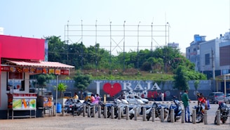 A bustling street scene features several parked scooters in front of a large billboard with the text 'I ❤️ RAJA'. The surroundings include a small street cafe with a red awning on the left, greenery and trees behind the billboard, and several people near the scooters. Modern buildings are visible in the background.