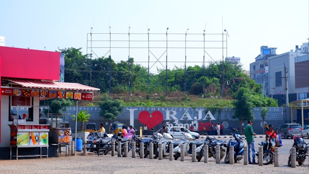 A bustling street scene features several parked scooters in front of a large billboard with the text 'I ❤️ RAJA'. The surroundings include a small street cafe with a red awning on the left, greenery and trees behind the billboard, and several people near the scooters. Modern buildings are visible in the background.