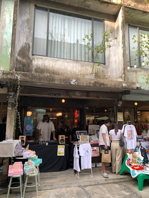 An outdoor market scene with a rustic building as the backdrop. A vendor stall is set up in front of a shop, displaying various items like T-shirts, books, and decorative items. People are browsing the items. A DJ is playing music from a table with speakers and a turntable. The setting conveys a casual, urban vibe with greenery and hanging lights adding to the ambiance.