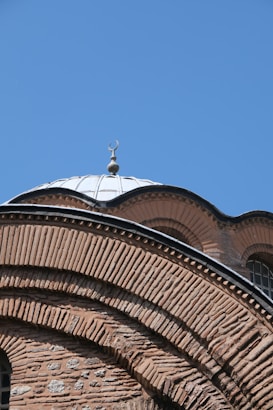 The image showcases a close-up view of an architectural structure featuring intricate brickwork and a prominent dome with a crescent-shaped ornament on top. The dome is made of metal, reflecting sunlight under a clear blue sky.