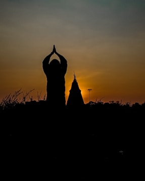 A serene silhouette of a person with hands pressed together above their head, suggesting a gesture of prayer or meditation. The background features a vibrant sunset sky with a gradient of orange, yellow, and blue hues, creating a peaceful and contemplative atmosphere. Additional silhouettes include structures and scattered foliage in the foreground.