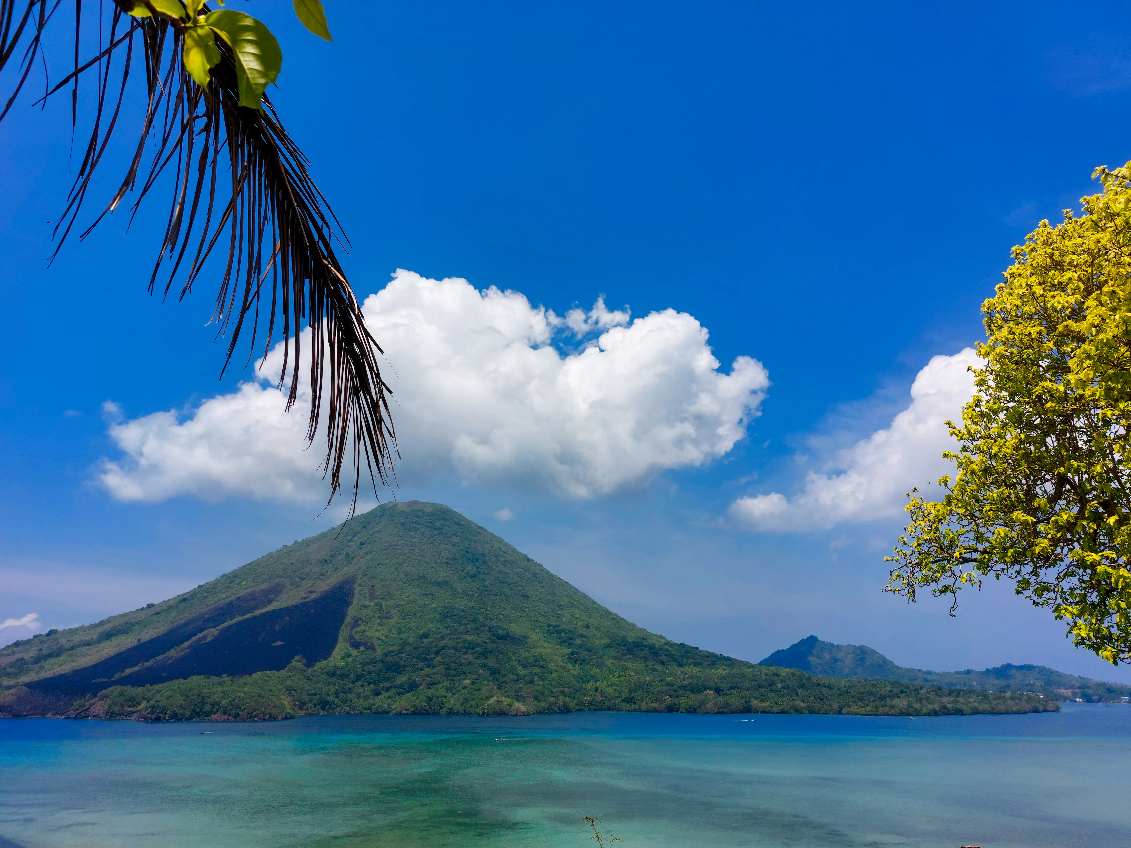 A cone-shaped volcano above turquoise water, framed by palm fronds in the Banda Islands