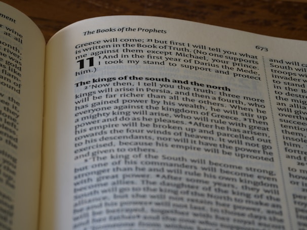 The image depicts an open book with a focus on a specific page. The text is clearly printed and includes a large bold number 11 at the start of a section, followed by a passage discussing historical events involving Greece. The pages are slightly curved, typical of an open book, and the background shows a wooden surface.