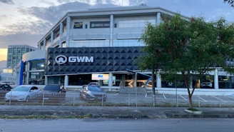 A modern multi-story building with a prominent black and white sign displaying 'GWM'. In front of the building, several parked cars are visible, lined up near a fence. A tree is situated to the right, next to the empty parking spaces. The sky is partly cloudy, and the lighting suggests it is either morning or late afternoon.