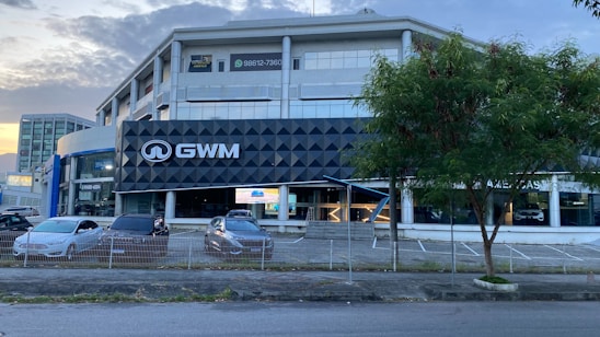 A modern multi-story building with a prominent black and white sign displaying 'GWM'. In front of the building, several parked cars are visible, lined up near a fence. A tree is situated to the right, next to the empty parking spaces. The sky is partly cloudy, and the lighting suggests it is either morning or late afternoon.