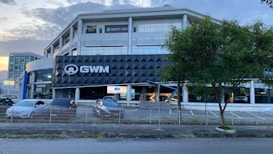 A modern multi-story building with a prominent black and white sign displaying 'GWM'. In front of the building, several parked cars are visible, lined up near a fence. A tree is situated to the right, next to the empty parking spaces. The sky is partly cloudy, and the lighting suggests it is either morning or late afternoon.