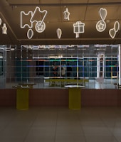 A mirrored wall reflects the image of two people standing inside a modern interior space. The ceiling is decorated with illuminated neon signs shaped like stars, hearts, tickets, and gift boxes. The floor is tiled and two yellow cylindrical tables are positioned against the mirrored wall.
