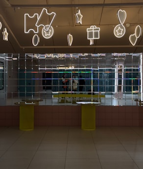 A mirrored wall reflects the image of two people standing inside a modern interior space. The ceiling is decorated with illuminated neon signs shaped like stars, hearts, tickets, and gift boxes. The floor is tiled and two yellow cylindrical tables are positioned against the mirrored wall.