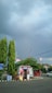 A small roadside kiosk is situated next to a parking area, surrounded by lush green trees. Overhead, the sky is overcast with dark gray clouds, suggesting impending rain. Several advertising posters and signs are seen around the kiosk, with a red and white color theme. A red metal fence runs alongside the street, adding to the urban environment.